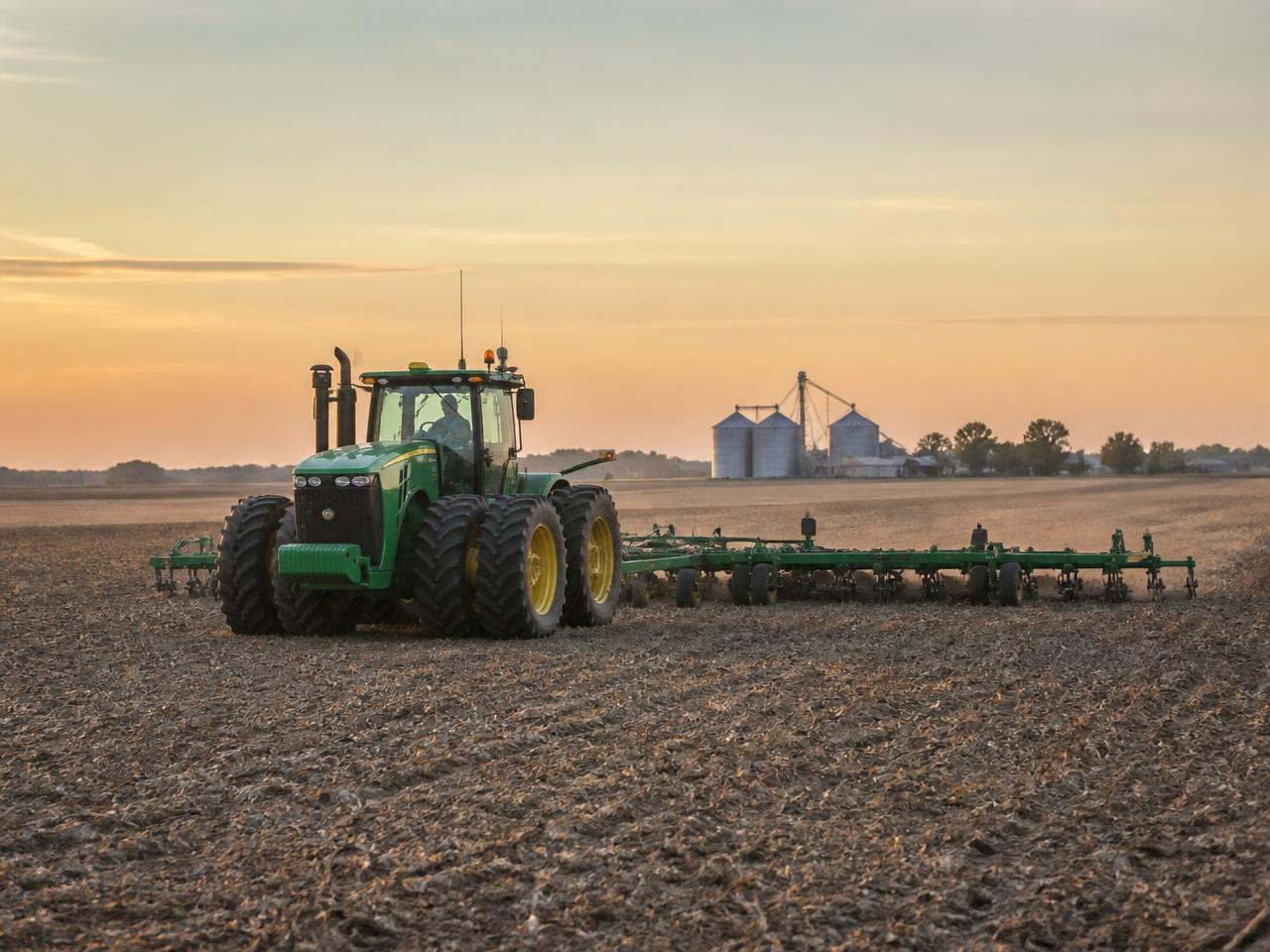 A tractor pulling a wide implement across a vast field at dawn with grain silos on the horizon
